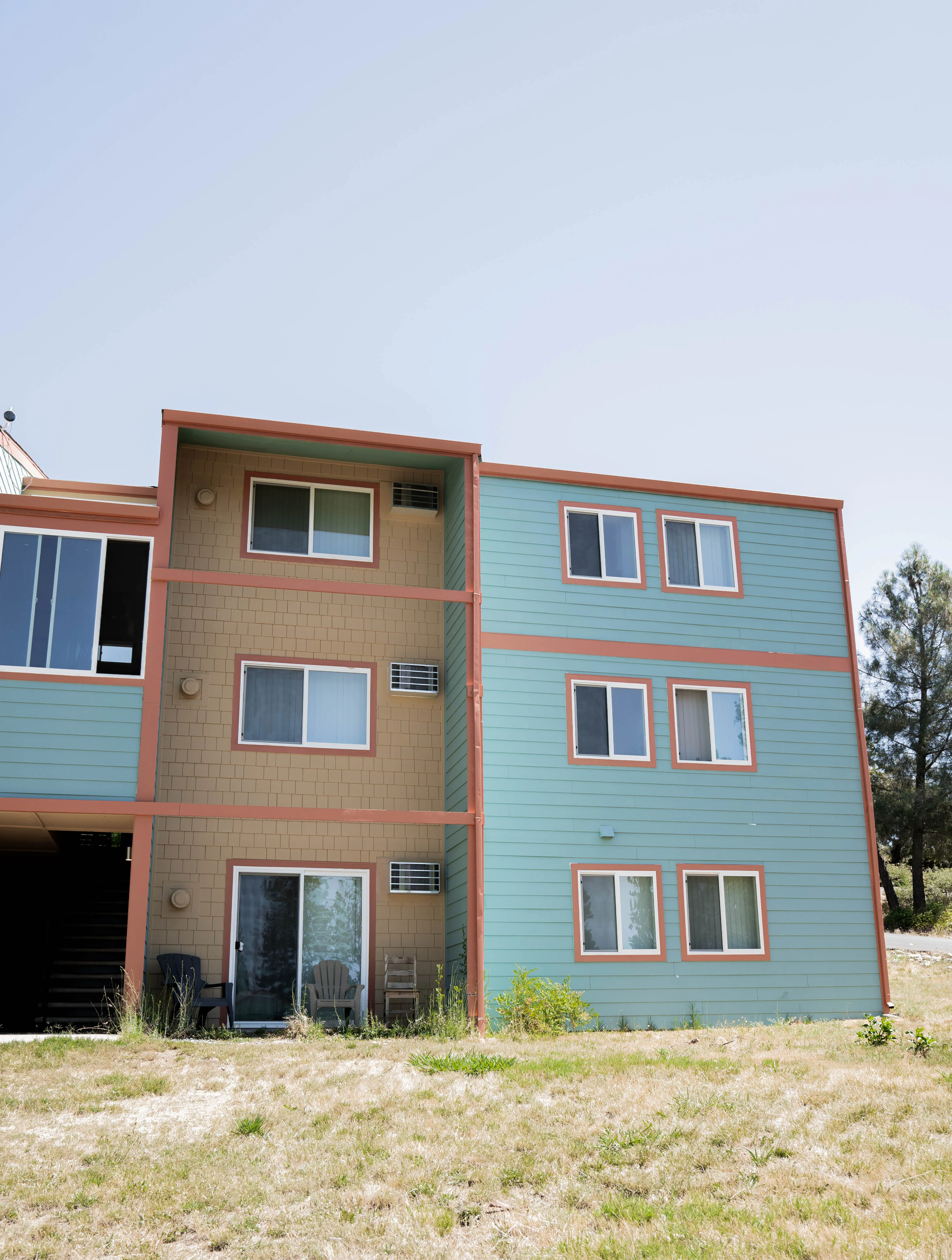 A multi-story residential building with blue and beige siding, featuring several windows and a sliding glass door, set against a clear sky.