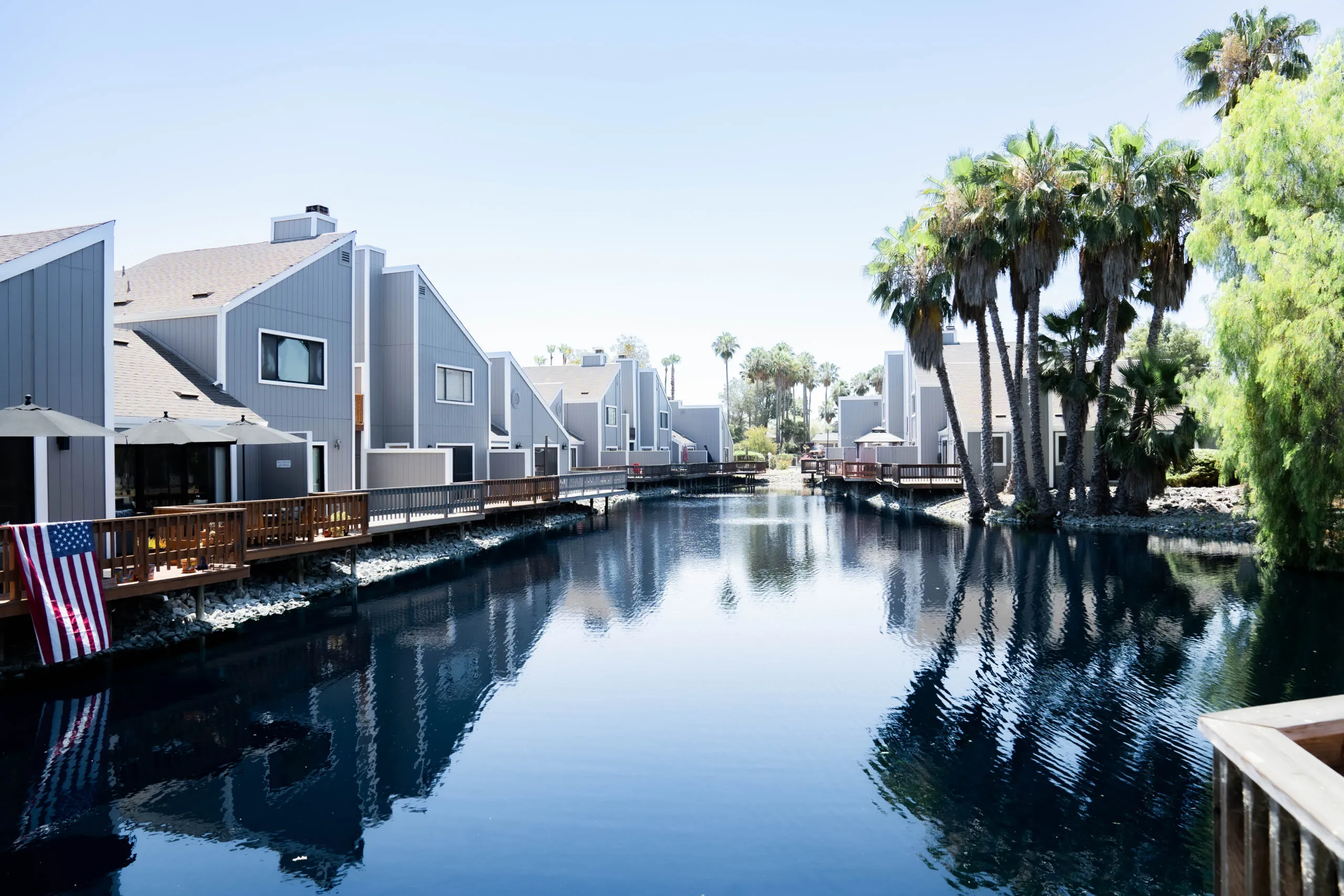 "Modern gray buildings with wooden decks and lush palm trees reflected in a tranquil waterway, designed by Heritage Exteriors Inc. in Sacramento."