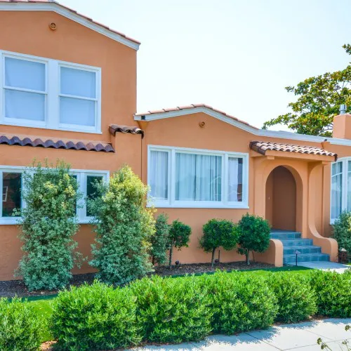 A renovated orange stucco house with white framed windows and a clay tile roof, showcasing a garden with shrubs, located in Berkeley.