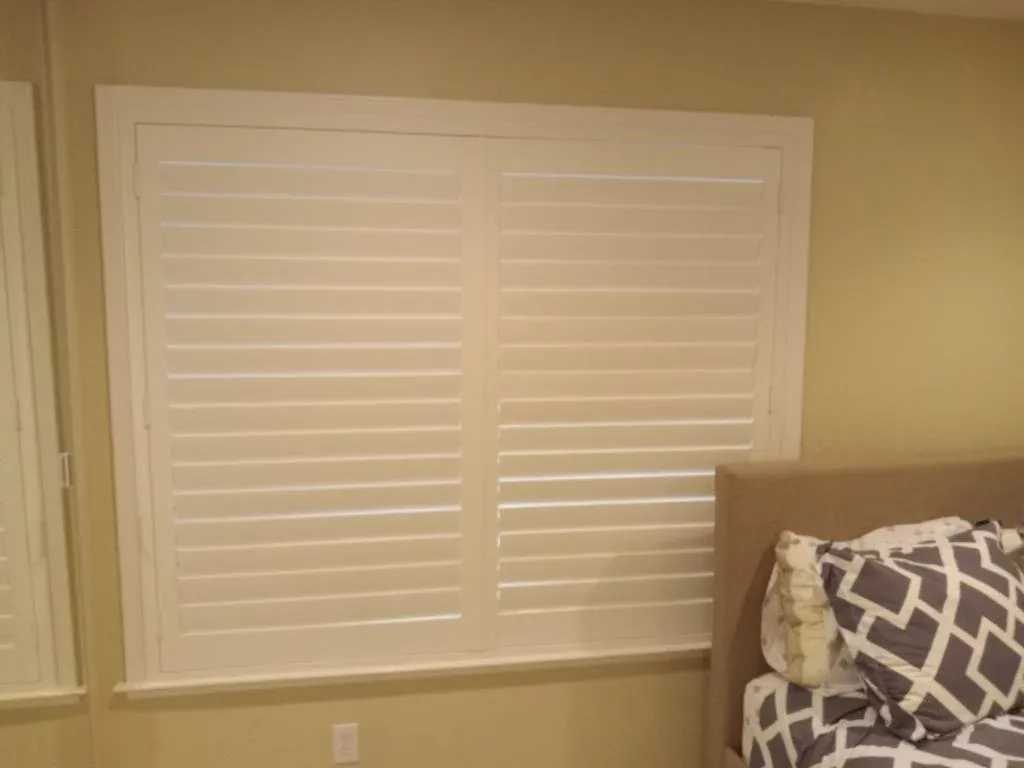 Closed white shutters on a window next to a bed with patterned pillows, emphasizing interior decor.