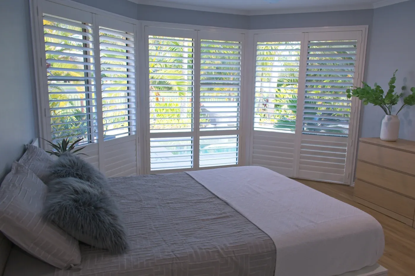 A bedroom featuring large windows with white shutters, a neatly made bed, and a dresser with a vase of greenery.