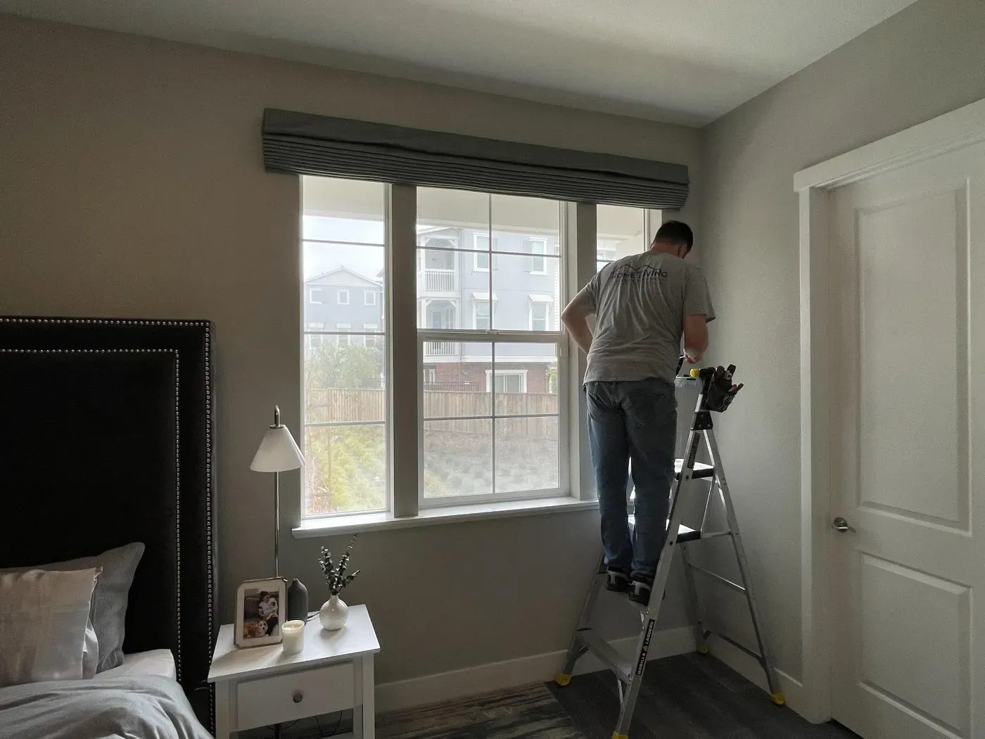 Person on a ladder installing a window treatment in a bedroom, with a bed and nightstand visible.