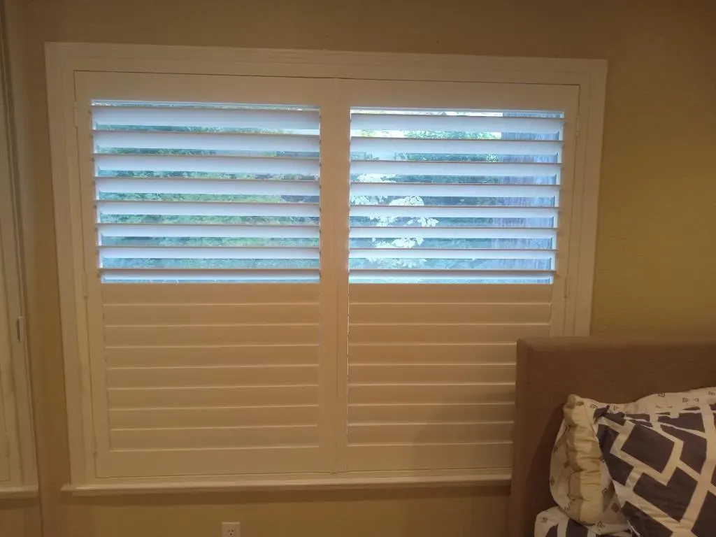 A window with white shutters partially open, allowing a view of greenery outside. The room has beige walls and a bed with patterned pillows is visible.