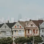 A row of colorful Victorian-style houses, showcasing intricate architectural details, against the backdrop of a city skyline.