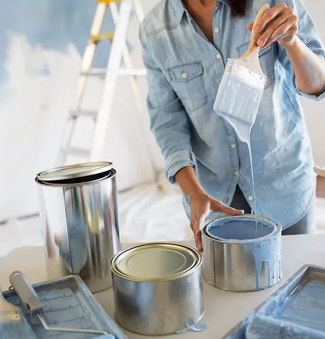 Person in denim shirt holding a brush with blue paint, surrounded by open paint cans and a ladder in the background.