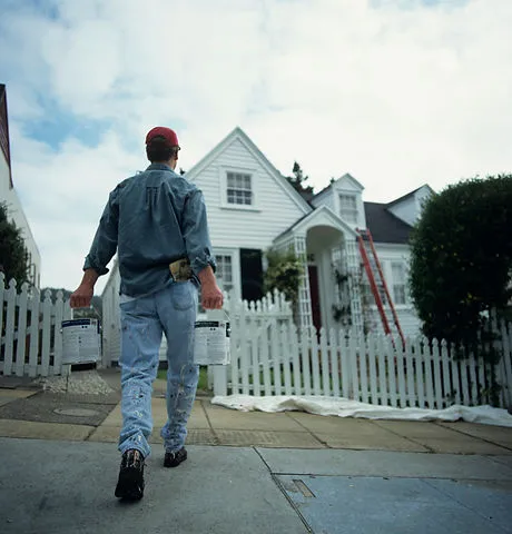 A painter carrying two paint cans approaches a white house with a picket fence and a ladder, symbolizing the professional services of Innovation Painting Plus in San Mateo.
