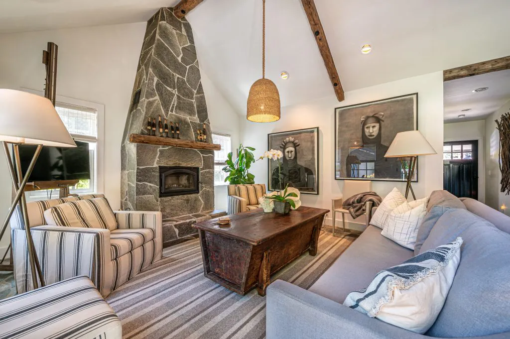 A cozy living room with a stone fireplace, striped armchairs, a wooden coffee table, and decorative art pieces, reflecting a blend of rustic and modern design.
