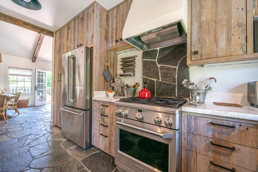 A kitchen featuring rustic wooden cabinets, a stainless steel refrigerator and stove, stone flooring, and a natural stone backsplash. Crafted by Intentional Contracting Inc. in Santa Rosa.