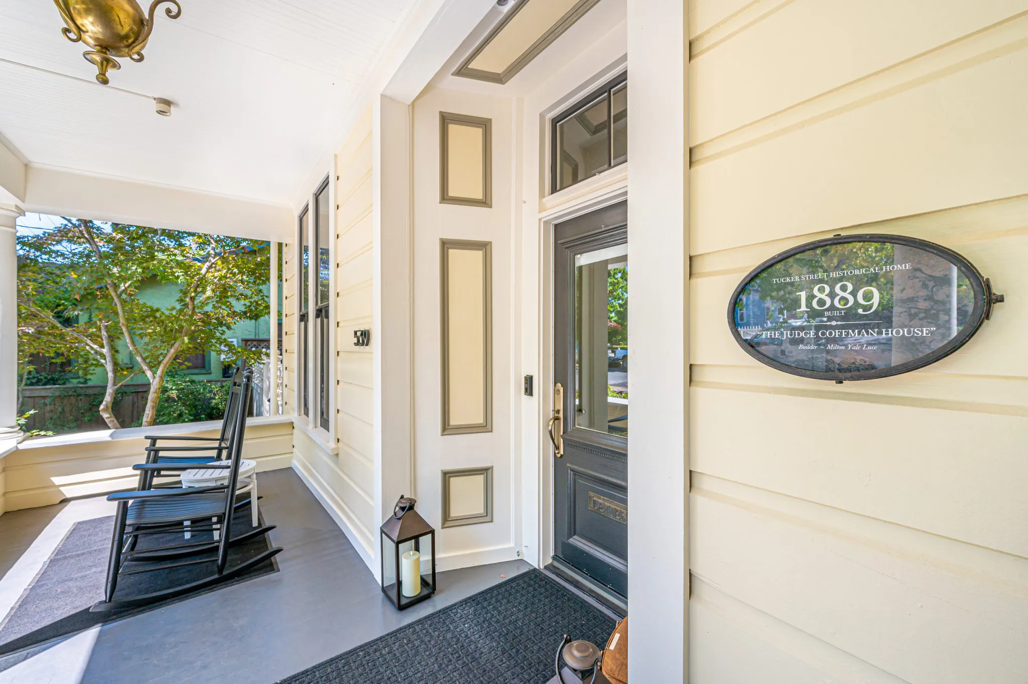 A porch view of a historical home featuring a sign reading "1889 The Judge Coffman House," with rocking chairs, lantern, and decorative paneling.
