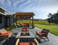 Outdoor seating area with modern wooden furniture and a fire pit under a pergola, set in a scenic landscape.