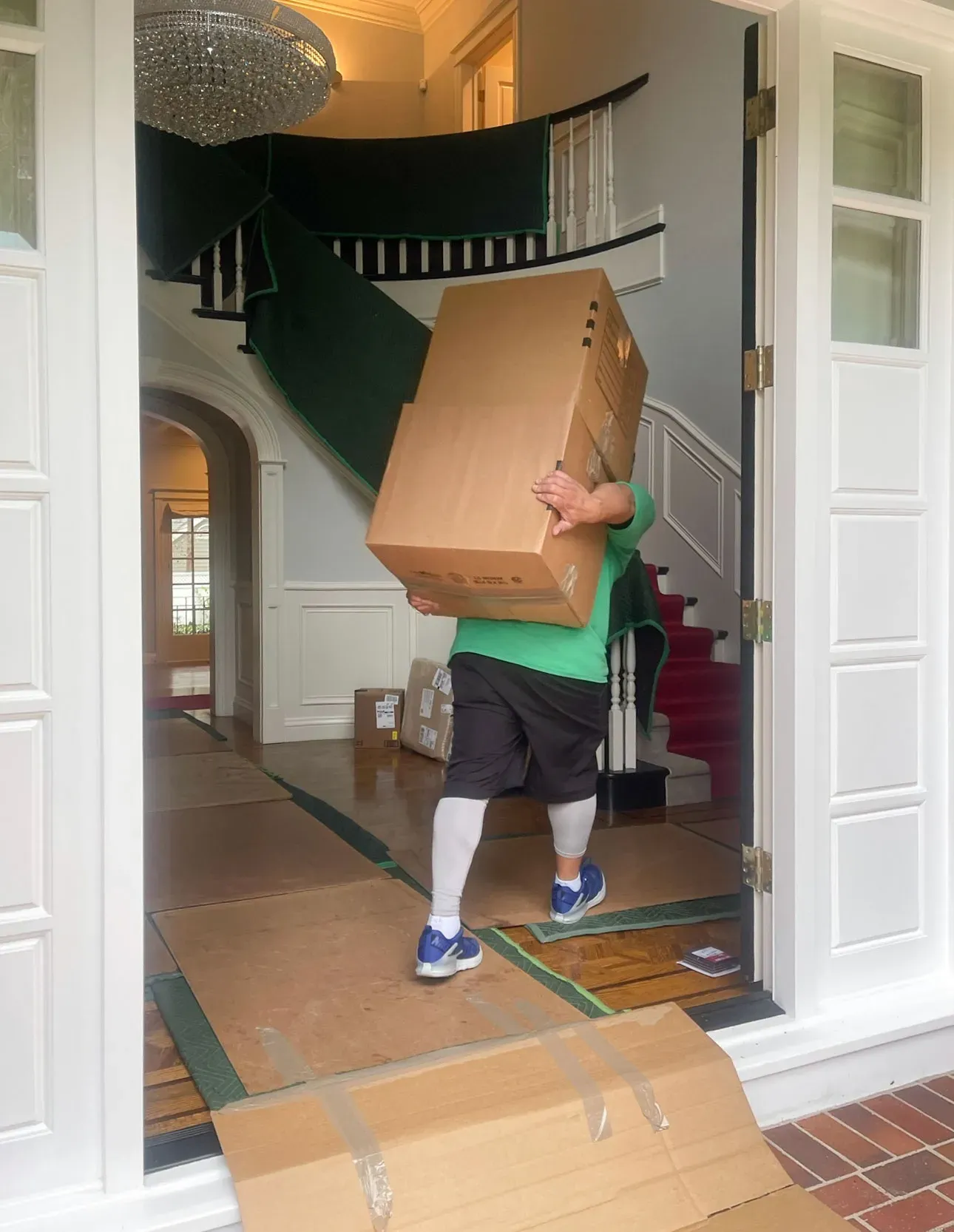 A mover in a green shirt and white leggings carries a large cardboard box into a home with a grand staircase in Walnut Creek. The floor is protected with cardboard sheets.