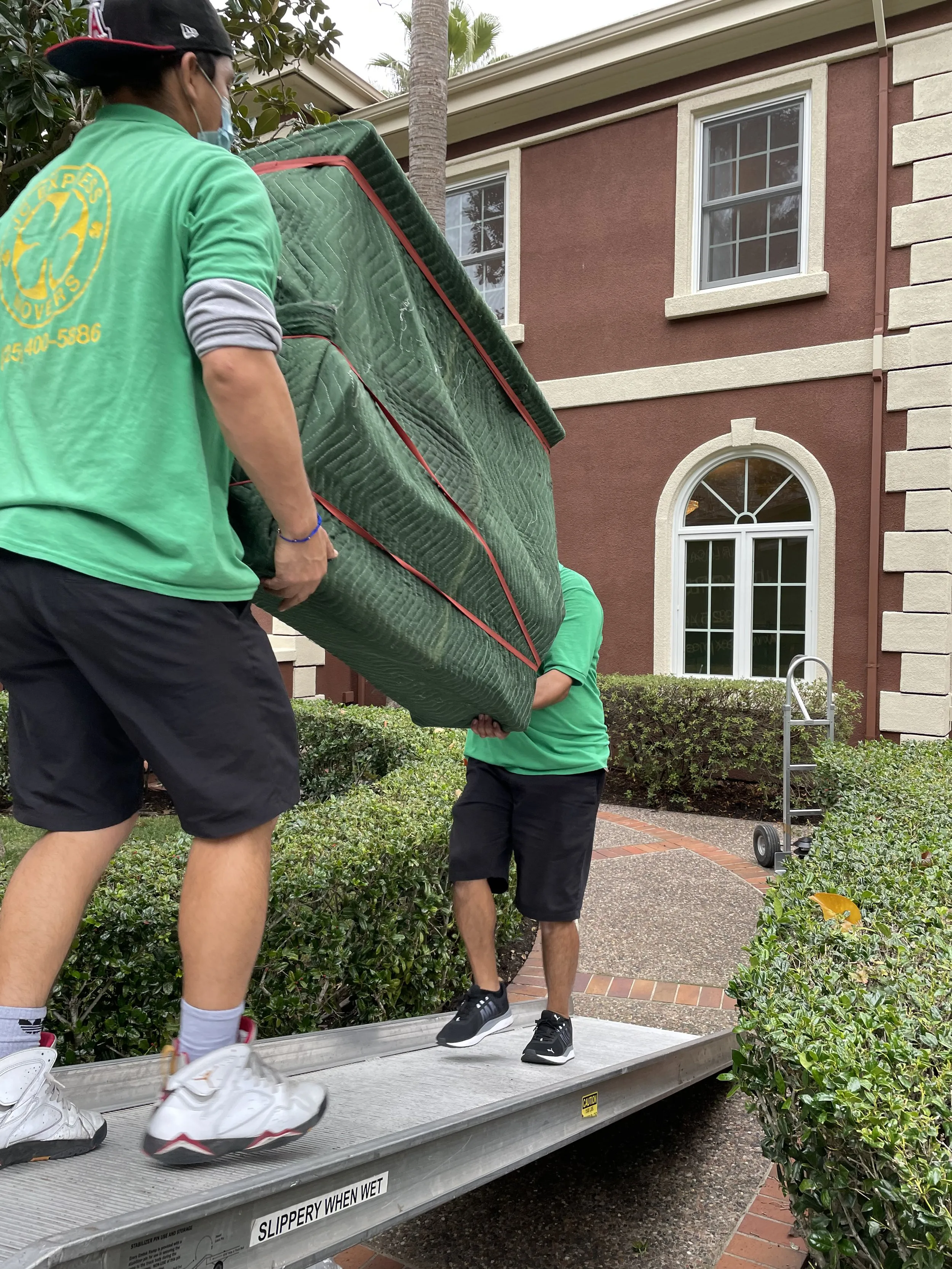 Two movers from JC Express Movers carry a large, wrapped item up a ramp outside a residential building in Walnut Creek, wearing green company shirts and using careful coordination.