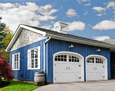 A blue garage with two white garage doors, adorned with stone accents under a bright blue sky.