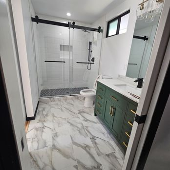 A modern bathroom featuring a glass-enclosed shower, marble floor tiles, green vanity with brass handles, and a spacious countertop.
