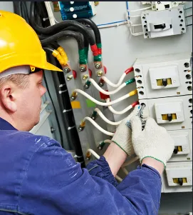 An electrician wearing a yellow helmet and gloves works on an electrical panel with various colored wires.