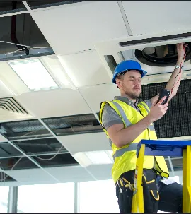 Electrician in a hard hat and reflective vest examines wiring on a ceiling light fixture while standing on a ladder.