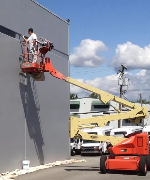 A worker from Joe Moore Painting & Roofing uses a lift to paint a large exterior wall in Windsor, surrounded by clouds and parked vehicles.