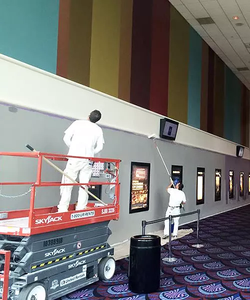 Two painters from Joe Moore Painting & Roofing working indoors in Windsor, using a lift and rollers to paint a high wall above framed posters.