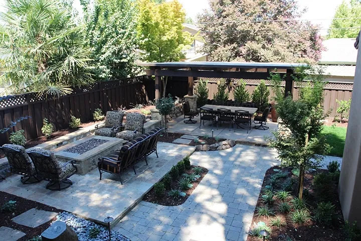 A landscaped backyard in Fremont designed by Koch & Associates, Inc., featuring a seating area with cushioned chairs around a fire pit, a pergola-covered dining table, and surrounding greenery.