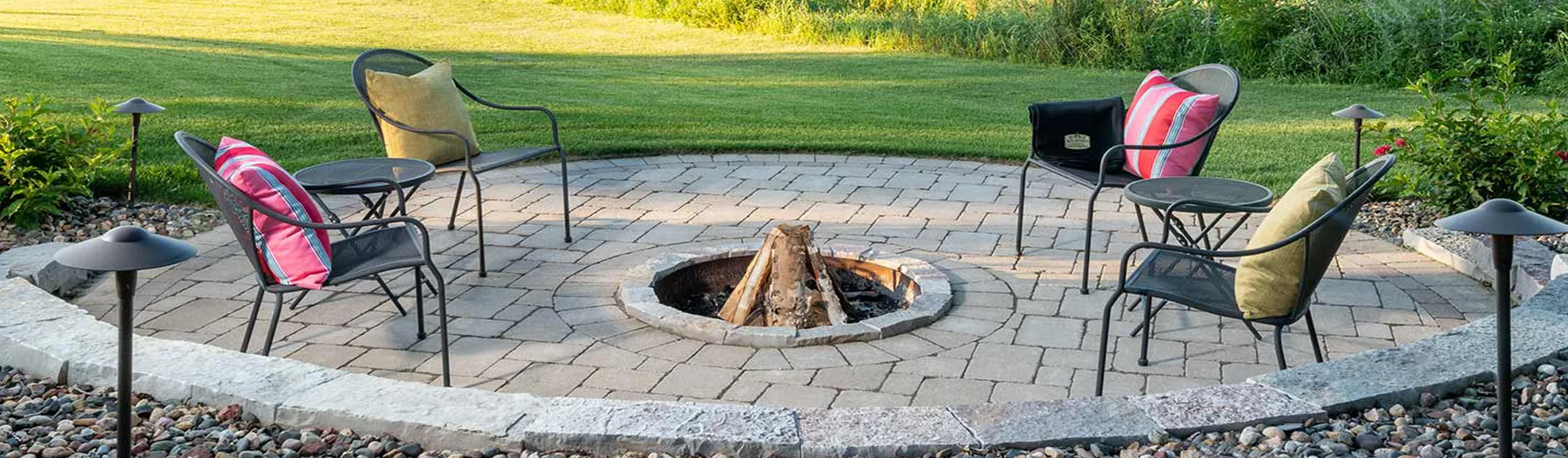 A round stone patio with a central fire pit, surrounded by outdoor chairs and a well-maintained lawn in bright sunlight.