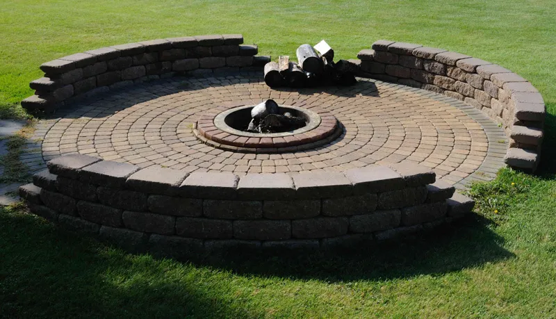 A circular stone fire pit designed with concentric brick layers surrounded by a low stone wall, set on a green lawn, created by Kodiak Construction in Martinez.