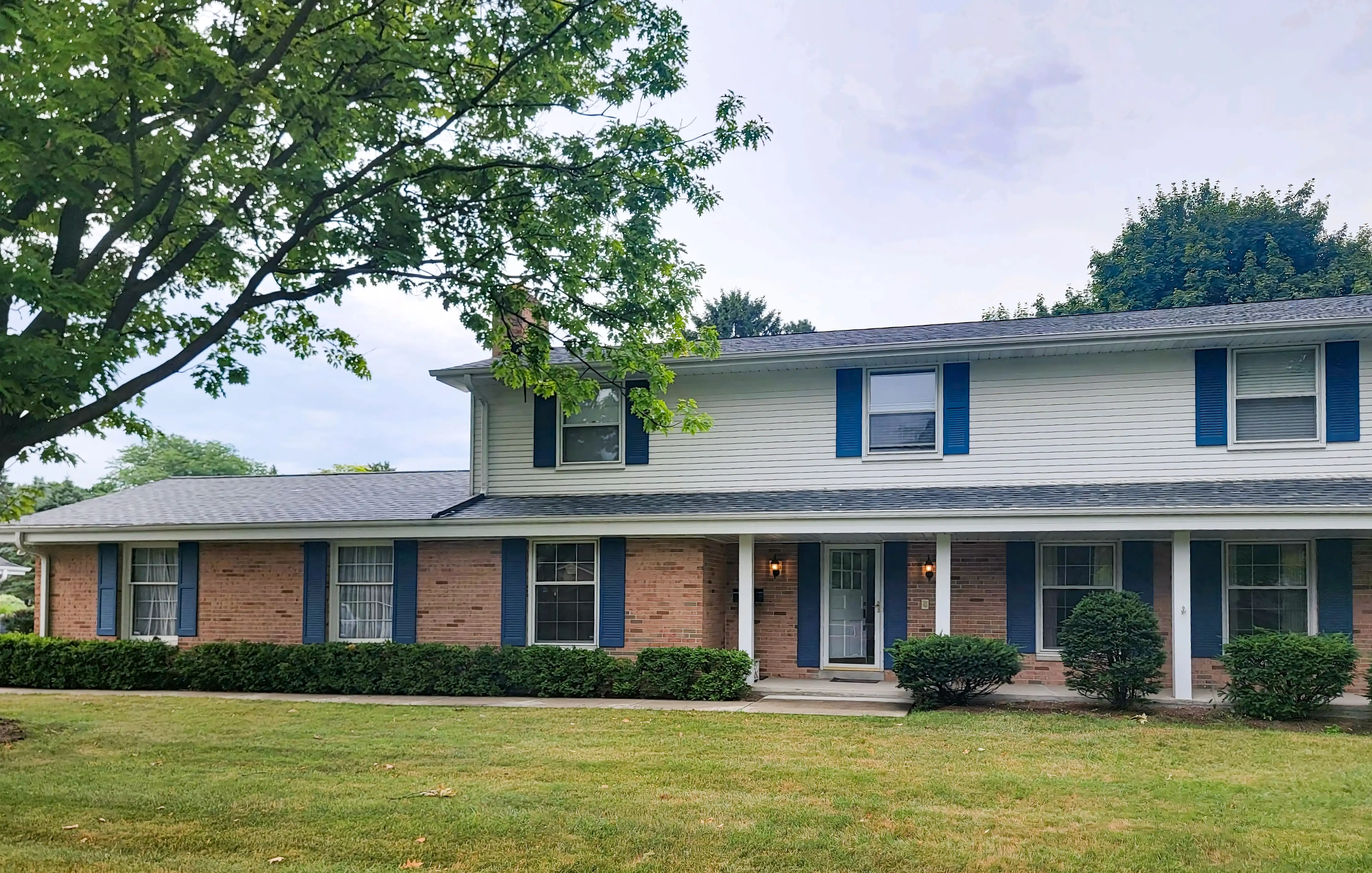A two-story suburban house with a brick facade, featuring white siding on the upper level and blue shutters, surrounded by a well-maintained lawn and trees.