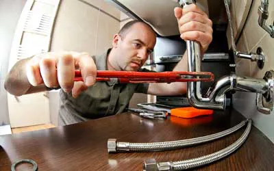 A professional plumber from LW Construction & Handyman Services using a wrench to adjust a pipe under a kitchen sink, with tools and hoses beside him.