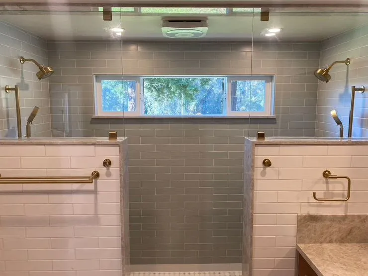 A modern dual shower with glass screens, gold fixtures, and a window in the center, designed by Labourdette Construction in Sebastopol.