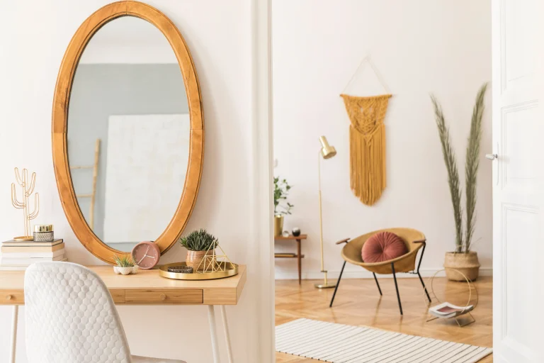 A contemporary interior view showing a chic workspace with a large oval mirror above a desk, and an adjoining room with a stylish chair, potted plants, and decorative wall hangings.