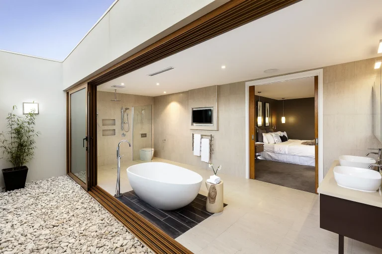 A modern bathroom with open sliding doors, featuring a freestanding bathtub, dual sinks, and a view into a bedroom.