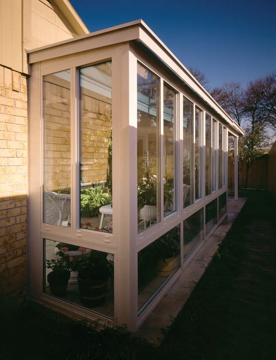 A modern sunroom with large glass windows attached to a brick house, featuring plants and wicker chairs inside.