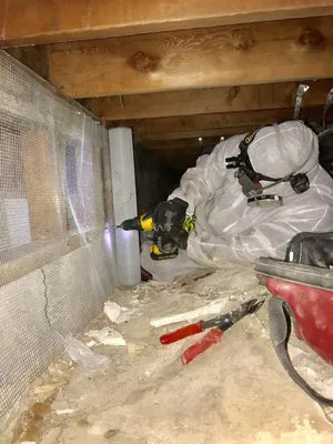 A technician in protective gear working in a crawl space, using a power tool to reinforce structural elements, with various tools scattered nearby.
