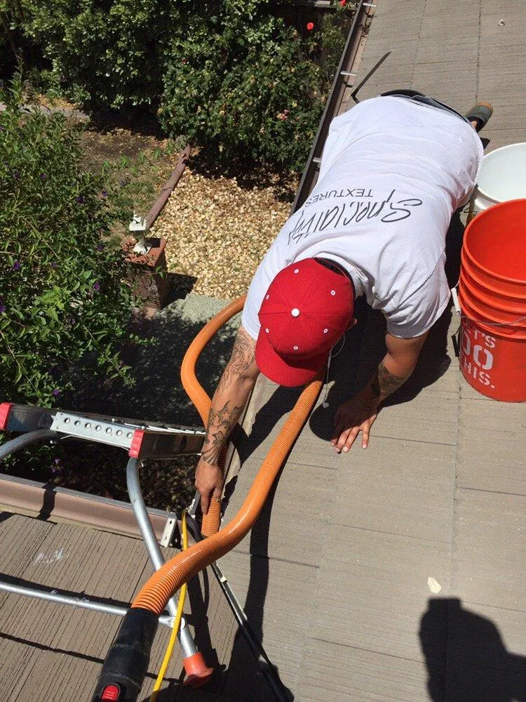 A person in a red cap cleaning gutters on a roof using a vacuum, with ladders and tools visible.