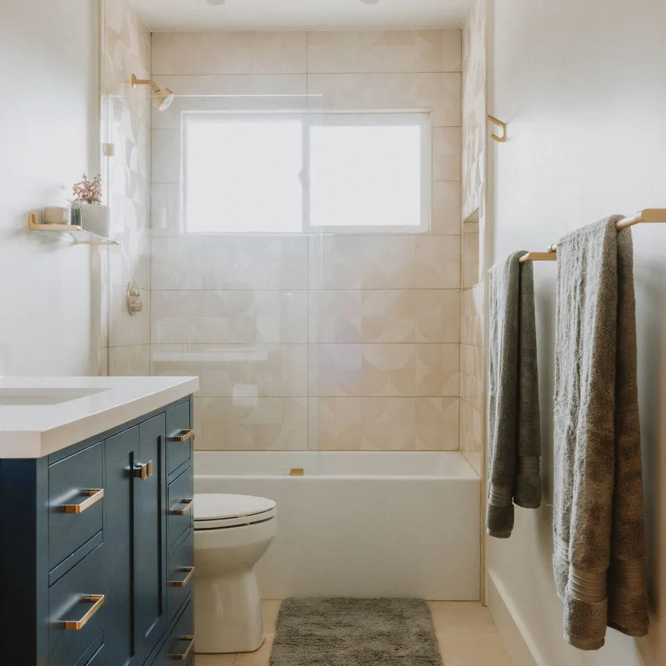 "A modern bathroom featuring a blue vanity, a bathtub, beige wall tiles, a window, and neatly hung gray towels."