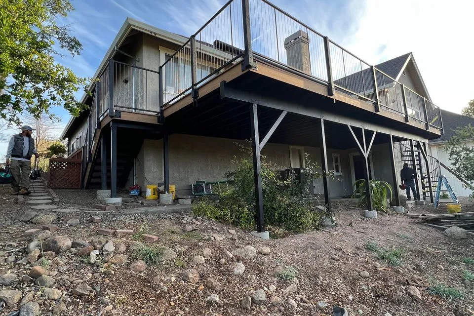 A large outdoor deck with metal railings and supporting beams, constructed by Make It Right Carpentry, Inc. in Novato. Two people are near the structure, indicating ongoing work.