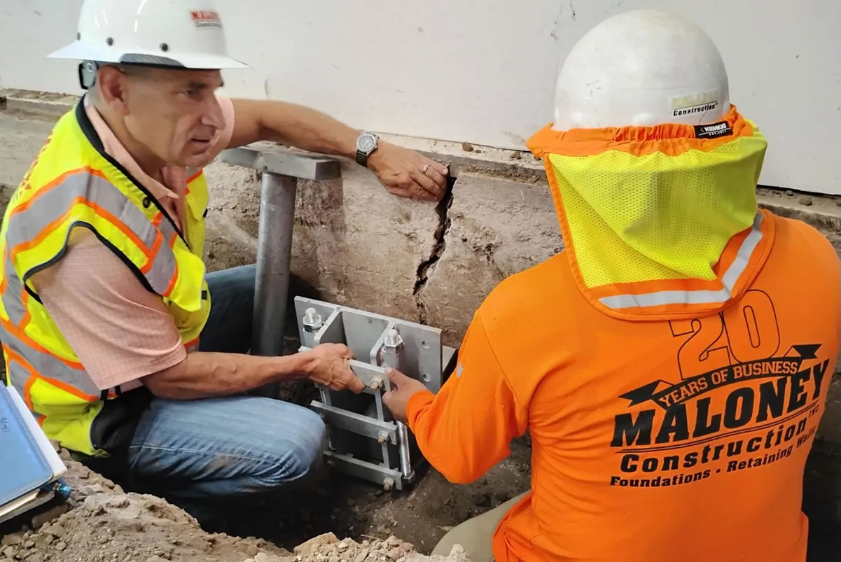 Two men from Maloney Construction, Inc. in Novato, wearing safety gear, work on reinforcing a building foundation with visible cracks.