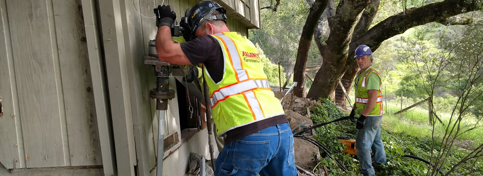 Two workers from Maloney Construction, Inc. in Novato performing foundation repair beside a building, surrounded by trees.