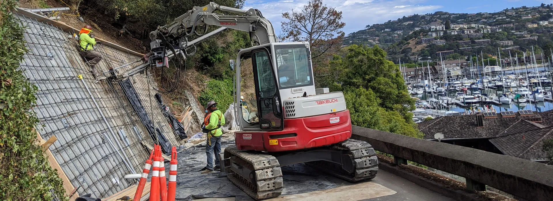 Construction workers using a Takeuchi excavator to reinforce a hillside with metal mesh in Novato, with a marina visible in the background.