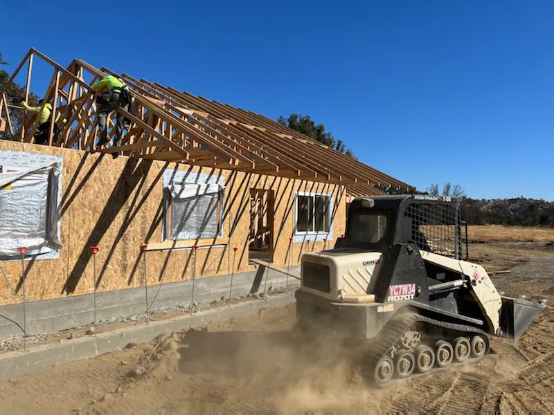 Construction site in Vallejo with a partially built wooden structure, workers on the roof, and a tracked loader moving dirt in the foreground.