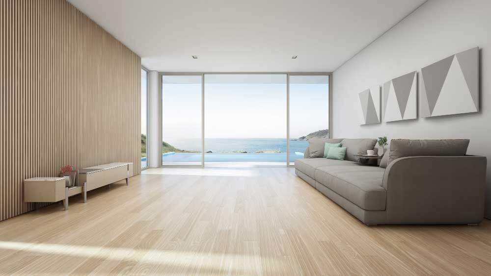 A modern living room with wooden flooring and large glass doors overlooking the ocean, featuring a grey sofa and minimalist decor.