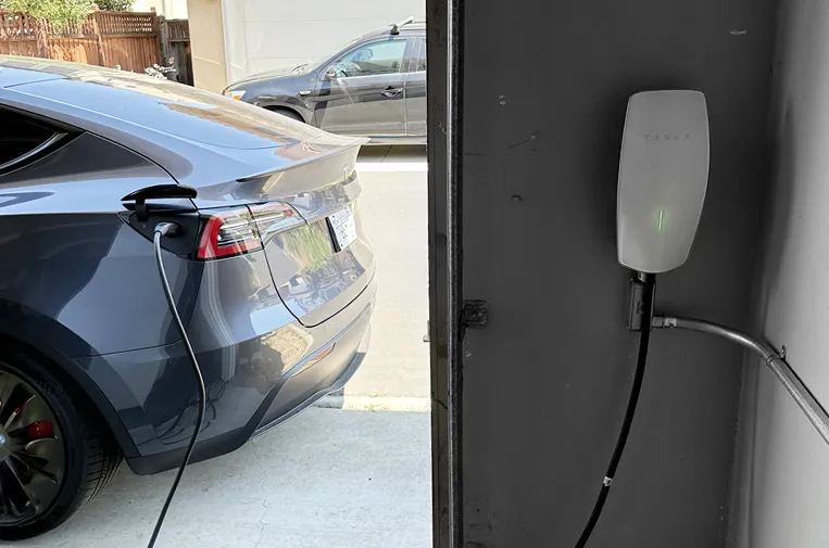 An electric vehicle is parked in a driveway, connected to a wall-mounted EV charging station, showcasing the charging setup.