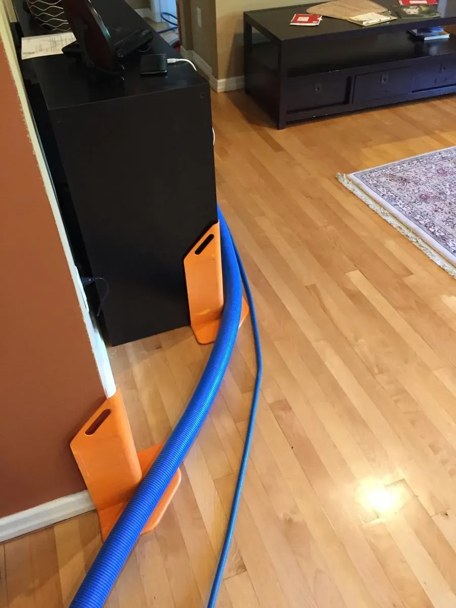 A blue hose from Maximum Carpet Cleaning equipment runs across a wooden floor, supported by orange stands to protect walls and furniture in a Pleasant Hill home.