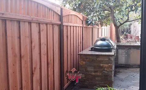 Wooden fence alongside a stone barbecue area with a grill, set in a backyard with trees, designed by Meza'sFence in San Jose.