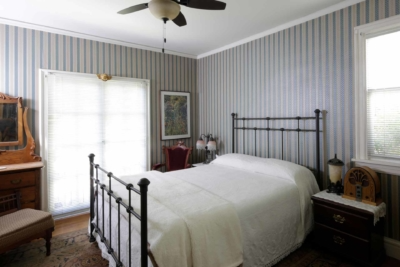A bedroom with striped wallpaper, featuring a metal bed frame, wooden furniture, and a ceiling fan.