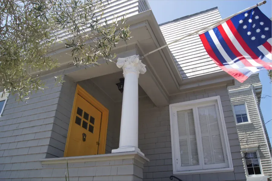 A close-up view of a house with gray siding and a prominent yellow door, featuring a white column and an American flag, painted by Michael Sack Painting, Inc. in San Francisco.