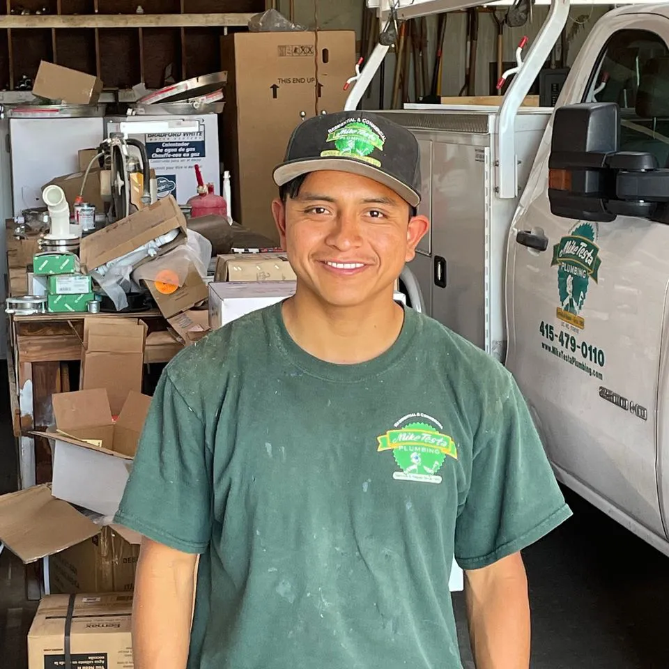 A smiling employee of Mike Testa Plumbing, Inc. in San Rafael stands in a workshop, with a truck and various plumbing tools and boxes visible in the background.