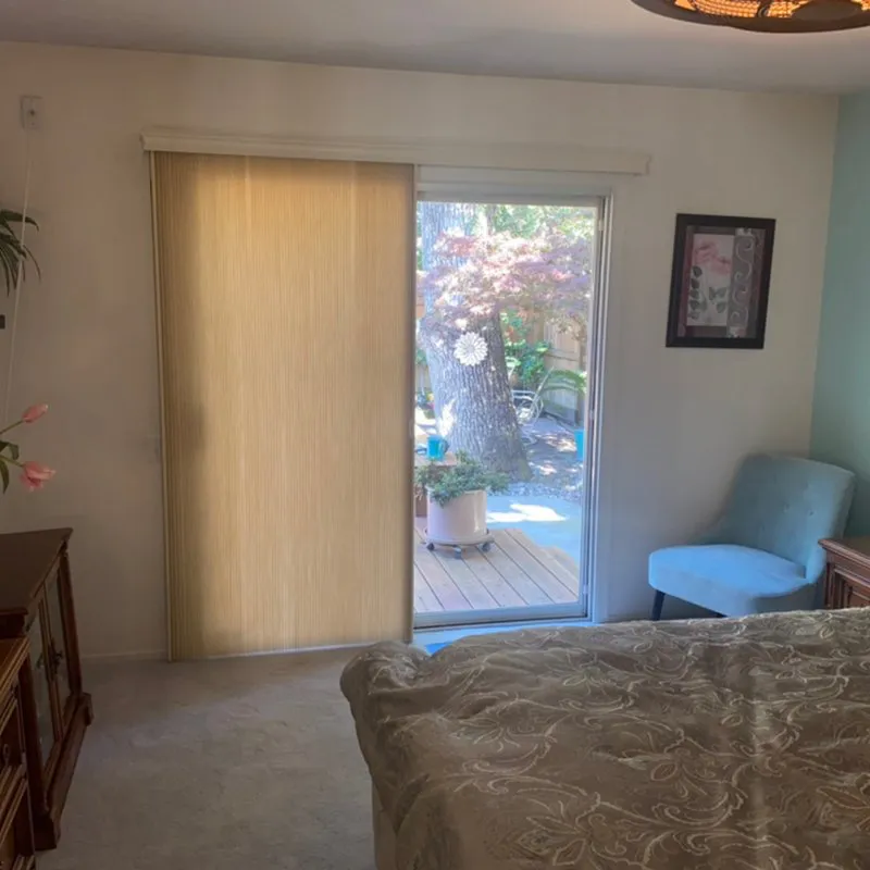 A cozy bedroom in Novato featuring a sliding glass door with vertical blinds by Miller Window Covering, offering a view of an inviting outdoor space.