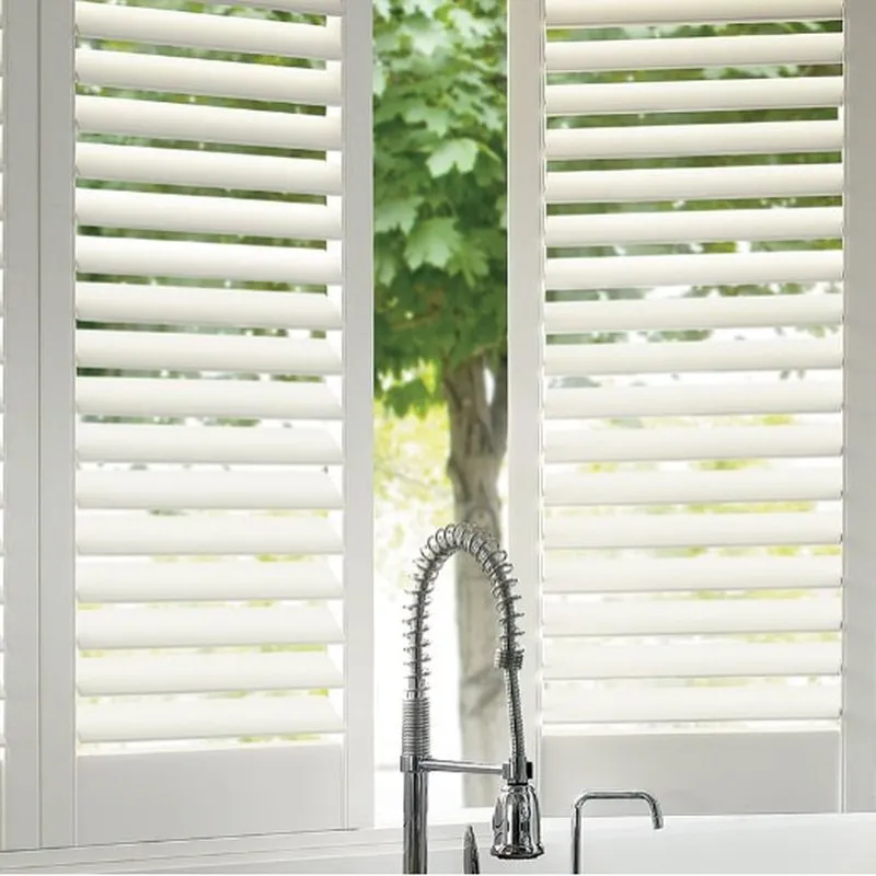 A kitchen with a modern faucet and white plantation shutters on the windows, allowing a view of a tree outside.