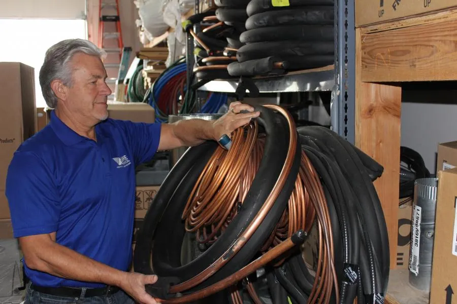A person in a blue Moore Mechanical Heating & Air Conditioning polo shirt inspects coiled copper tubing and hoses in a storage area.