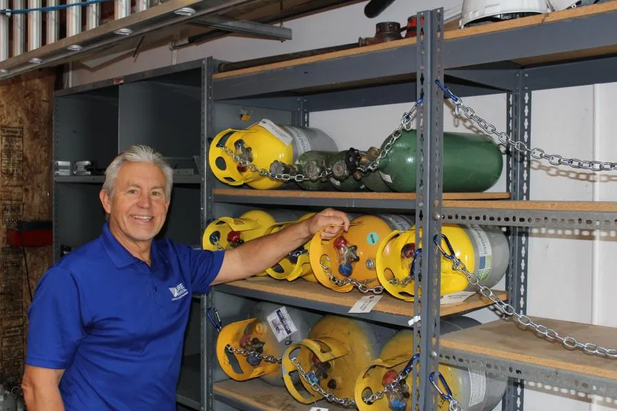 A person in a blue shirt stands near shelves stocked with secured yellow and green gas cylinders, showcasing the organized storage at Moore Mechanical Heating & Air Conditioning in Dublin.
